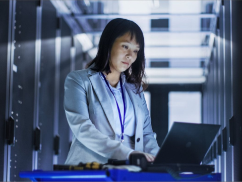 A female IT Professional works on her laptop in the server room.